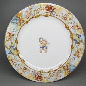May include: A white ceramic plate with a decorative border featuring cherubs in various poses, with a central cherub figure. The border has gold, blue, and yellow accents. The plate is likely for decorative or display purposes.