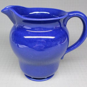 May include: A cobalt blue ceramic pitcher with a curved handle and a spout. The pitcher has a rounded body and a glossy finish. The handle is arched, and the spout is designed for easy pouring. This kitchenware item is suitable for serving beverages.