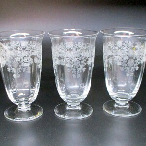 May include: Three clear glass goblets with a floral etched design. The goblets have a flared rim, a slender body, and a round base. The etched design is visible on the upper portion of the glass. The goblets are arranged in a row against a dark background.