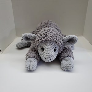 May include: A gray crocheted stuffed sheep toy with a fluffy texture. The sheep is lying down with its head resting on the ground.