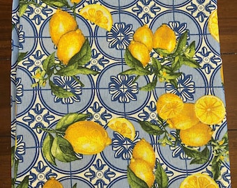 May include: A folded blue and white tablecloth with a repeating pattern of lemons and green leaves. The lemons are yellow and the leaves are green. The background has a blue and white tile design.