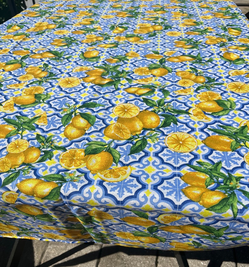 May include: A white tablecloth with a blue and yellow lemon pattern. The tablecloth features a repeating pattern of lemons, lemon slices, and green leaves on a blue and white tiled background.