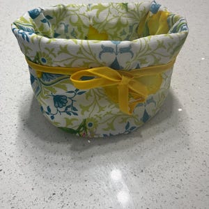 May include: A fabric basket with a floral pattern in shades of green, yellow, and blue. The basket has a yellow ribbon tied around the middle. The interior of the basket is visible.