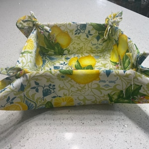 Puede incluir: Una cesta de tela plegable con un diseño de estampado de limón. La cesta está hecha de una tela blanca con hojas verdes y limones amarillos. La cesta está plegada y lista para usar.