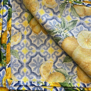 May include: A folded blue and white fabric with a lemon pattern. The fabric has a yellow border and a white background with blue and yellow geometric designs. The lemons are yellow with green leaves.