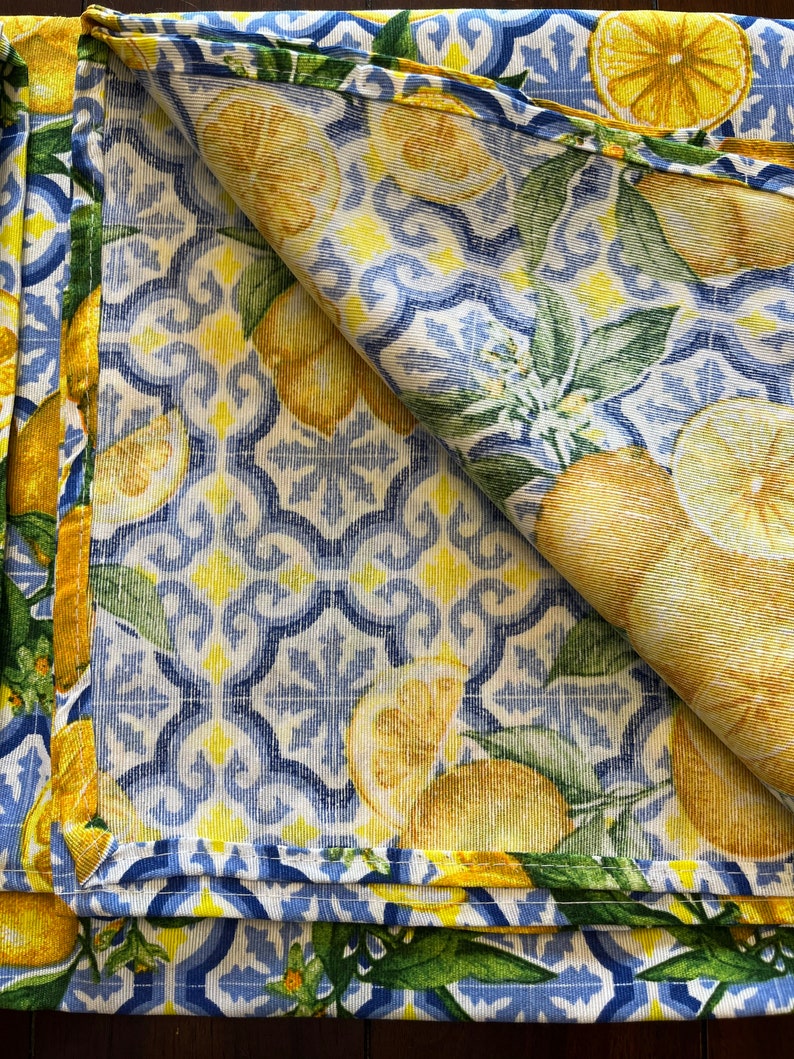 May include: A folded blue and white fabric with a lemon pattern. The fabric has a yellow border and a white background with blue and yellow geometric designs. The lemons are yellow with green leaves.