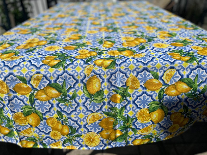 May include: A white tablecloth with a blue and yellow lemon pattern. The tablecloth features a repeating pattern of lemons, leaves, and blue and yellow tiles.