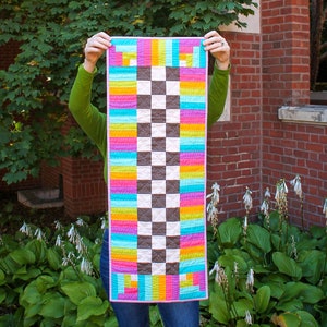 May include: A colorful, handmade quilt with a checkerboard pattern in the center and vibrant stripes of turquoise, pink, yellow, and orange. The quilt is held up in front of a brick building and green foliage.