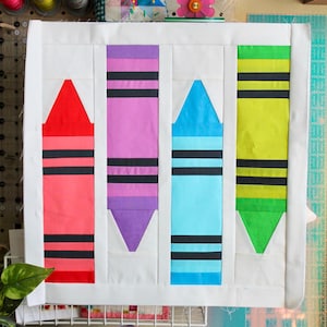 Könnte beinhalten: Ein weißes Stoffpanel mit vier bunten Crayon-Blöcken. Die Crayons sind rot, lila, blau und grün. Das Stoffpanel hängt an einem weißen Drahtgestell. Eine Box mit dem Text "RUBY+BEE SWATCH SET" befindet sich im Vordergrund.