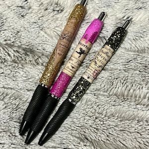 May include: Three glitter pens with black grips. One pen is gold with "Felia Felicis" text, another is pink with "morte" text, and the third is black with "Polyjuci Potion" text. The pens are on a fluffy, gray surface.