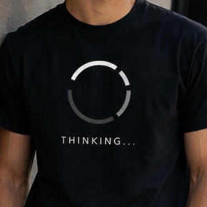 Könnte beinhalten: Ein schwarzes T-Shirt mit kurzen Ärmeln und einer weißen, kreisförmigen Lade-Grafik in der Mitte der Brust. Unter dem kreisförmigen Symbol ist das Wort "THINKING..." in einer klaren, weißen, serifenlosen Schriftart aufgedruckt.