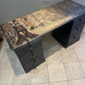 May include: Black desk with a steampunk design. The desktop features a detailed image of a woman in steampunk attire, gears, and the word "SHEZZ". The desk has multiple drawers and brass knobs.