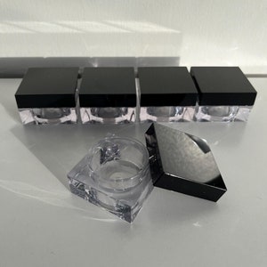Peut inclure: Cinq contenants carrés transparents avec des couvercles noirs. Un contenant est ouvert et le couvercle est posé à côté.