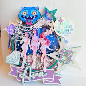 May include: A layered birthday cake topper featuring three stylized figures in white outfits, a blue cat, musical notes, and a disco ball. The design includes the name "Ava" and the number "13" in a star shape, with glitter accents.