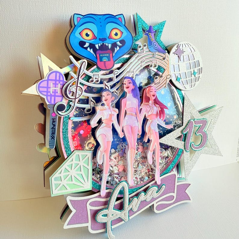 May include: A multi-layered, glittery cake topper featuring three stylized figures, musical notes, a disco ball, and a blue cat face. The name "Ava" is displayed on a banner, with the number "13" on a star.