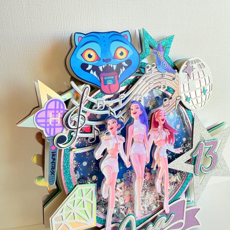 May include: A layered paper art piece featuring a blue cat face, musical notes, and three stylized figures in white outfits. The design includes a disco ball, stars, and the number 13, with glitter accents. The overall theme is a birthday celebration.