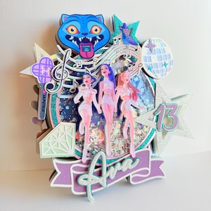 May include: A multi-layered birthday cake topper featuring three stylized figures, musical notes, a disco ball, and a blue cat face. The topper includes the name "Ava" and the number "13" in a purple and teal color scheme.