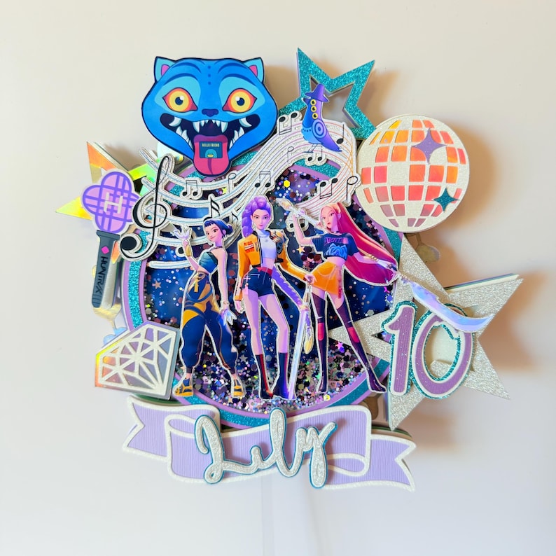 May include: A layered cake topper featuring three animated figures, a blue cat, musical notes, and a disco ball. The topper includes the number 10 and the word "July" in script. The color scheme includes blue, purple, orange, and silver glitter accents.