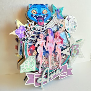 May include: A multi-layered, glittery cake topper with a blue cat face, musical notes, and three figures. The topper includes the name "Ava" and the number "13" in a star shape. The overall design is colorful and celebratory.