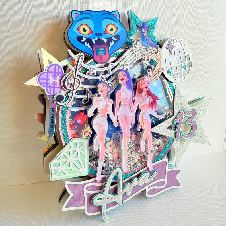 May include: A multi-layered, glittery cake topper with a blue cat face, musical notes, and three figures. The topper includes the name "Ava" and the number "13" in a star shape. The overall design is colorful and celebratory.