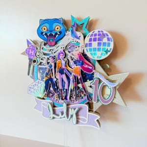 May include: A layered cake topper featuring a blue cartoon cat, a disco ball, and three stylized figures. The design includes musical notes, stars, and the word "July" on a banner. The number "10" is also visible.