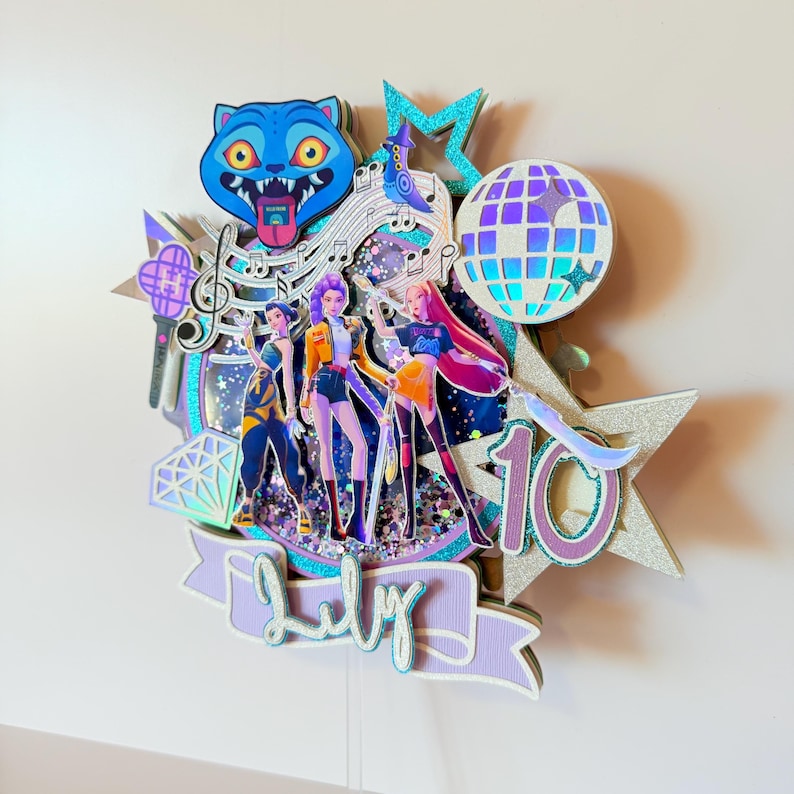 May include: A layered cake topper featuring a blue cartoon cat, a disco ball, and three stylized figures. The design includes musical notes, stars, and the word "July" on a banner. The number "10" is also visible.
