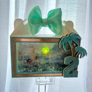 May include: A white box with a teal bow on top, featuring a framed scene with a tropical theme. The frame has a copper glitter border. Palm trees and the number 2 are on the side. The scene has a glowing light.