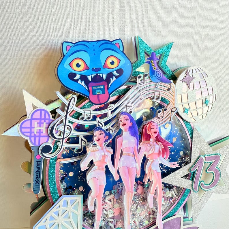 May include: A layered birthday cake topper featuring three stylized figures, musical notes, and a blue cat face. The design includes a disco ball, a star with the number 13, and a holographic background with glitter accents.