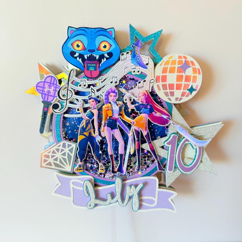 May include: A layered cake topper featuring three stylized figures, musical notes, a disco ball, and a blue cat with its tongue out. The topper includes the word "July" on a banner and the number "10".