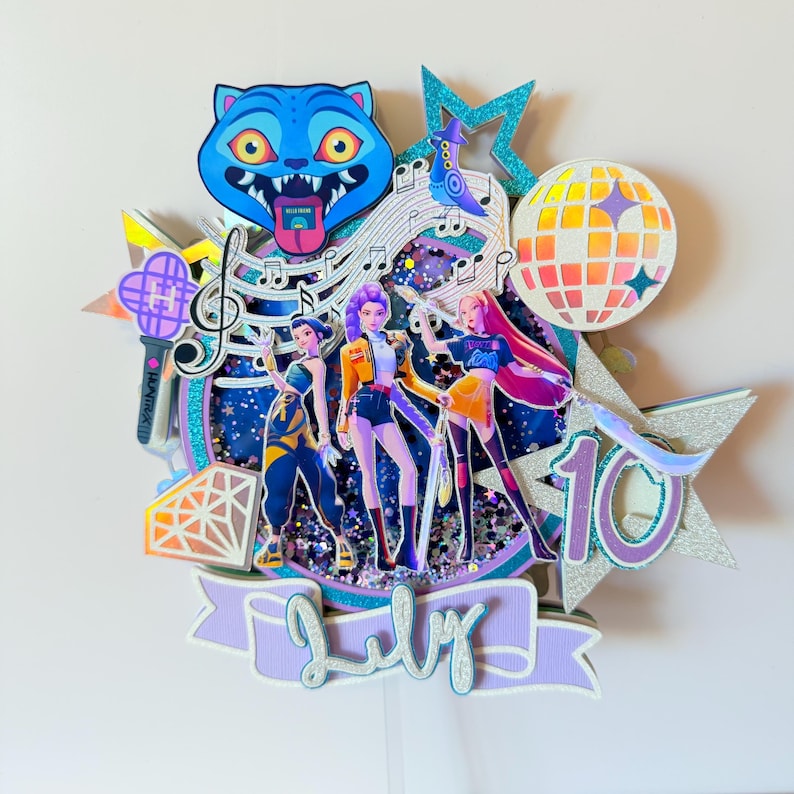 May include: A multi-layered cake topper featuring a blue cat, musical notes, a disco ball, and three stylized figures. The topper includes the number 10 and the month of July. The design incorporates glitter and various colors, creating a festive and celebratory theme.
