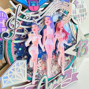 May include: A layered, glittery cake topper featuring three stylized figures in white outfits, holding hands. Musical notes, a disco ball, and the number 13 are also included. The background has a blue and silver glitter effect. The word "Ara" is visible.