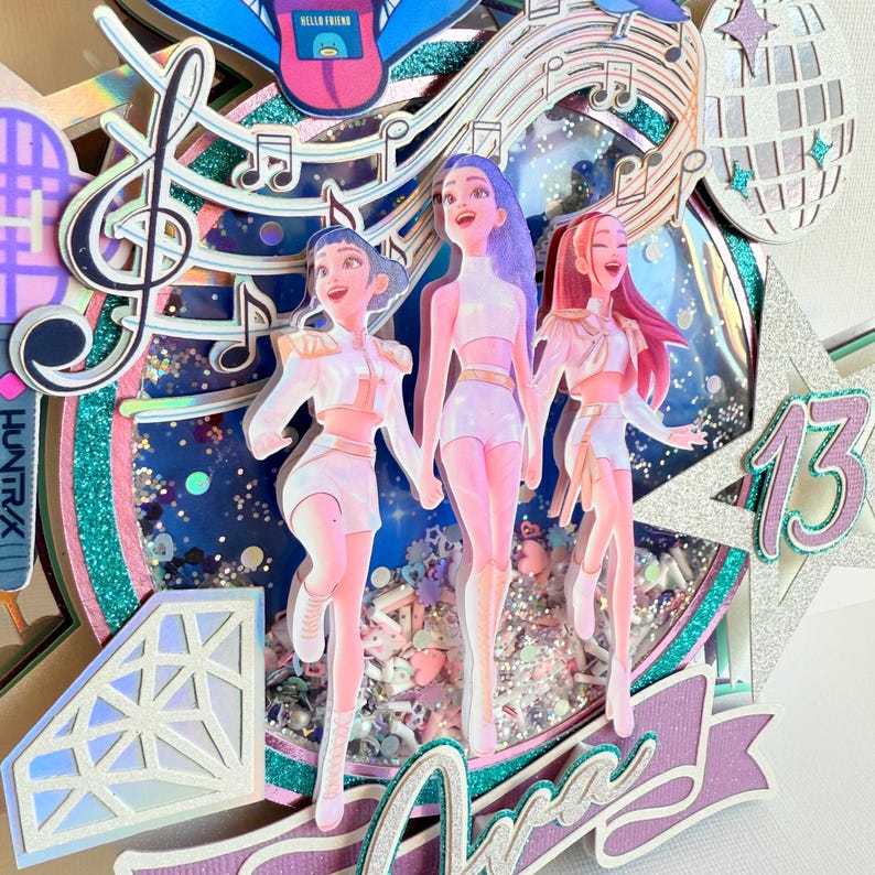 May include: A layered, glittery cake topper featuring three stylized figures in white outfits, holding hands. Musical notes, a disco ball, and the number 13 are also included. The background has a blue and silver glitter effect. The word "Ara" is visible.