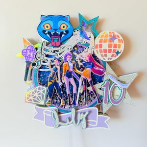 May include: A layered cake topper featuring three animated figures, a blue cat, musical notes, and a disco ball. The topper includes the word "July" and the number "10" in a decorative font. The color scheme includes blue, purple, orange, and silver.
