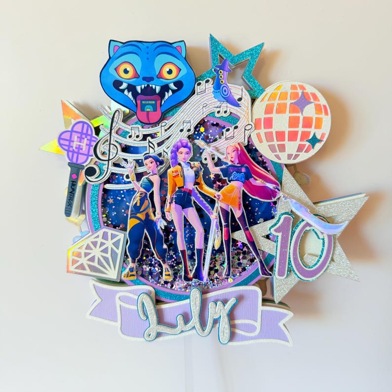 May include: A layered cake topper featuring three animated figures, a blue cat, musical notes, and a disco ball. The topper includes the word "July" and the number "10" in a decorative font. The color scheme includes blue, purple, orange, and silver.