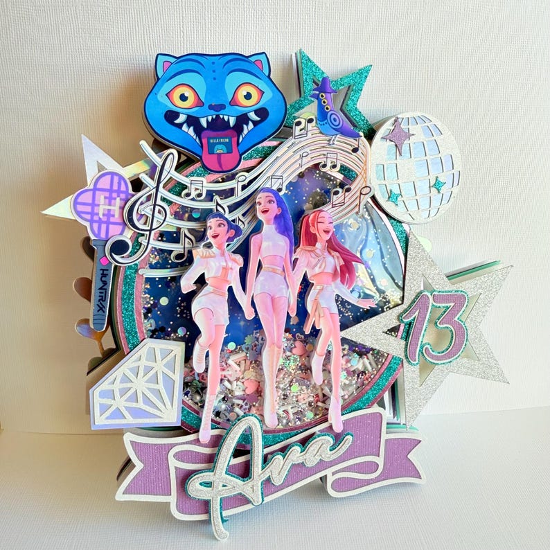 May include: A multi-layered cake topper featuring three stylized figures, musical notes, and a disco ball. The topper includes a blue cat, a star with the number 13, and a banner with the name "Ava". The color scheme includes shades of blue, pink, and purple.