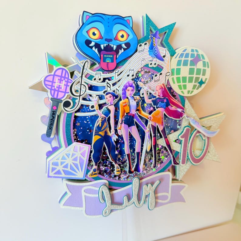 May include: A layered cake topper featuring three animated figures with colorful outfits, musical notes, and a disco ball. A blue cat-like character is at the top. The text "July" is on a banner, and the number "10" is also present.