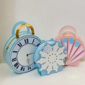 Puede incluir: Tres cajas de regalo de fiesta de papel azul y blanco con forma de reloj, copo de nieve y concha. El reloj tiene detalles dorados y números romanos. El copo de nieve tiene un diseño de copo de nieve blanco. La concha tiene un diseño iridiscente rosa y blanco.