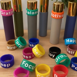 May include: A set of colorful silicone rings with words like "Gratitude", "Balance", "Motivate", "Forgive", "Inspired", "Energy", "Focus", "Sleep", "Happy", "Self", "Ground", "Joy", "Cheer", "Pass", and "Wake" printed on them. The rings are used for essential oil bottles.