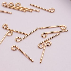 Puede incluir: Postes de pendientes dorados con diseño de bucle. Los postes son rectos con una pequeña parte trasera plana para asegurar a la oreja. Varias piezas están dispersas sobre una superficie de color claro.