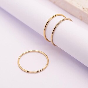 Può includere: Tre delicati anelli d'oro. Due anelli sono impilati su una superficie bianca, mentre il terzo anello è posizionato separatamente. Gli anelli hanno un design sottile e minimalista e lo sfondo è di un colore neutro.