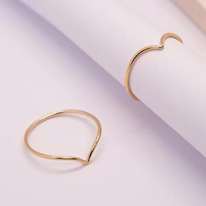 Pode incluir: Dois delicados anéis dourados. Um anel apresenta um design em forma de V, o outro tem uma forma curva de meia-lua. Os anéis são exibidos em uma superfície branca, destacando seu design minimalista e elegante. Um acessório simples, mas elegante.