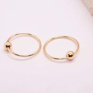 Könnte beinhalten: Zwei goldfarbene Nasenringe mit einem kleinen Kugelakzent. Die Ringe sind kreisförmig und haben eine glatte, polierte Oberfläche. Der Schmuck ist vor einem weichen, hellen Hintergrund platziert.