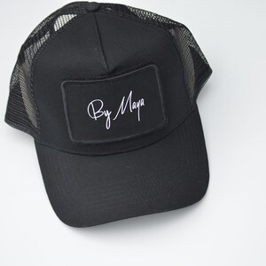 May include: Black trucker hat with a black patch that says "By Maya" in white cursive writing.
