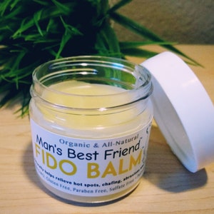 May include: A clear glass jar with a white lid containing a yellow balm. The jar has a label that reads "Man's Best Friend Organic & All-Natural FIDO BALM" and states that the balm helps relieve hot spots, chafing, and abrasions. The label also says that the balm is gluten free, paraben free, and sulfate free.