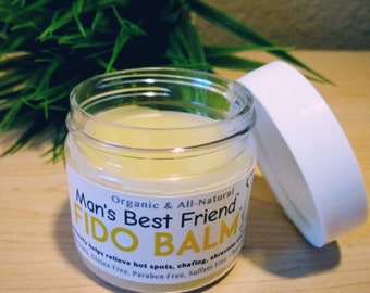 May include: A clear glass jar with a white lid containing a yellow balm. The jar has a label that reads "Man's Best Friend Organic & All-Natural FIDO BALM" and states that the balm helps relieve hot spots, chafing, and abrasions. The label also says that the balm is gluten free, paraben free, and sulfate free.
