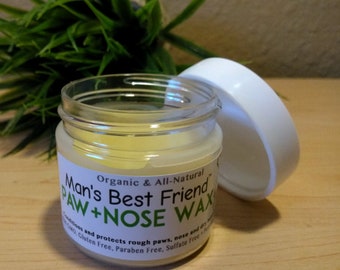 May include: A clear glass jar with a white lid, containing a yellow wax. The label reads "Man's Best Friend PAW + NOSE WAX" with "Organic & All-Natural" above. The product is designed to condition and protect paws and noses. The jar is open, and the background includes green foliage.