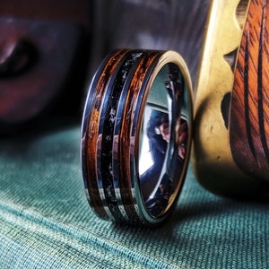 May include: A close-up of a silver ring with a wood and black inlay design. The ring is set against a backdrop of wood and a green surface. The word "BURL" is at the bottom of the image.