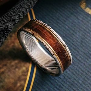Puede incluir: Primer plano de un anillo de madera y plata con una incrustación de madera marrón y una banda de plata con un patrón trenzado. El anillo está sobre una superficie azul.