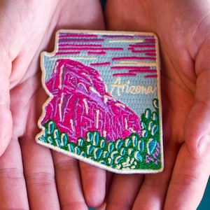 May include: Embroidered patch in the shape of Arizona, featuring a pink mountain scene, blue sky, and green cacti. The word "Arizona" is embroidered in white. The patch is held in open hands.