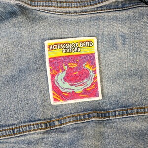 May include: A rectangular embroidered patch featuring a colorful landscape of Horseshoe Bend, Arizona, with the text HORSESHOE BEND ARIZONA in white letters. The patch displays shades of pink, purple, yellow, and light blue, attached to blue denim.